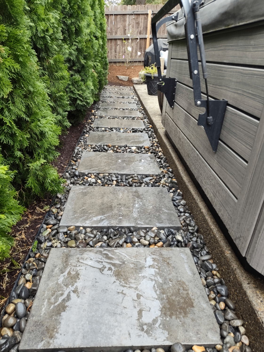 Modern stepping stone pathway with river rock beside a backyard spa and cedar hedge